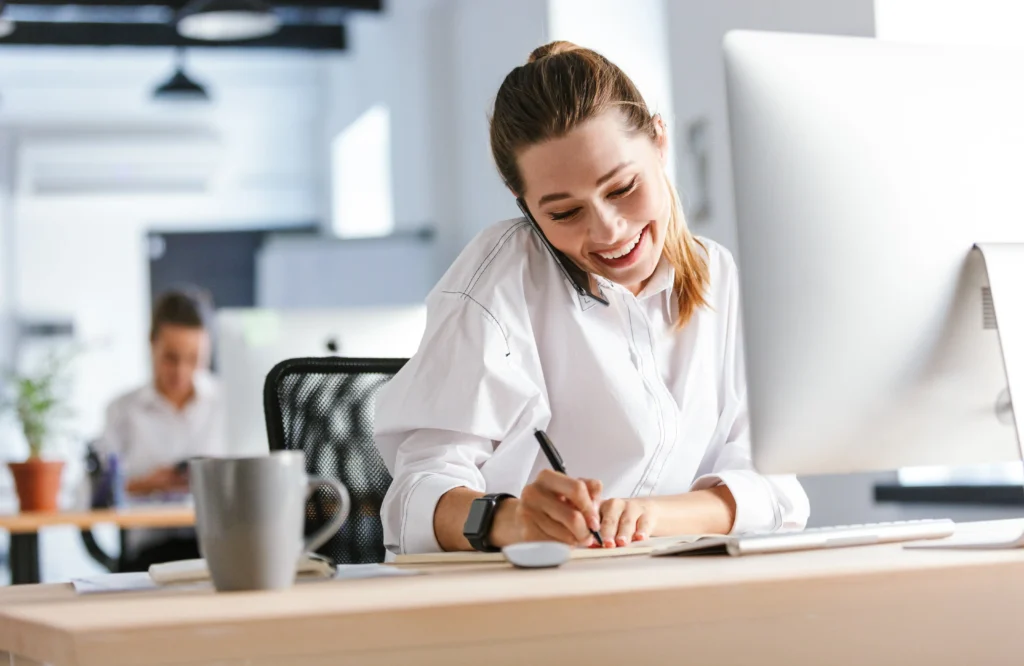 Lächelnde Mitarbeiterin am Schreibtisch telefoniert und macht sich Notizen im Büro
